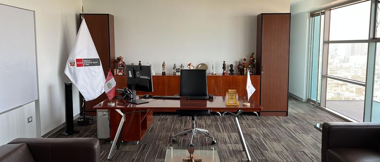 Despacho institucional con escritorio de madera, credenza con reconocimientos y banderas del Perú